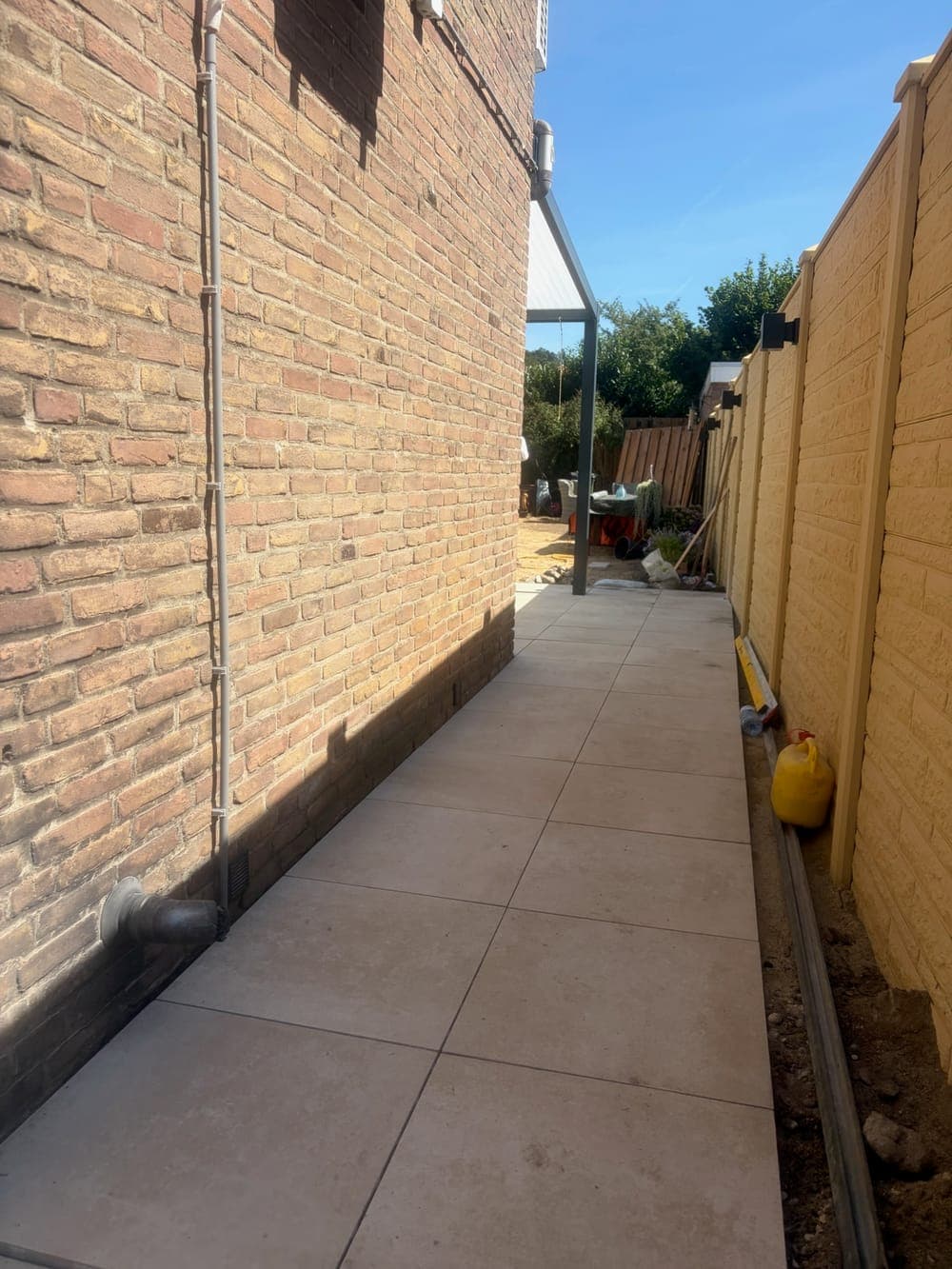 Het smalle pad van betontegels langs de gevel en de schutting, strak uitgelijnd tot achter in de tuin.