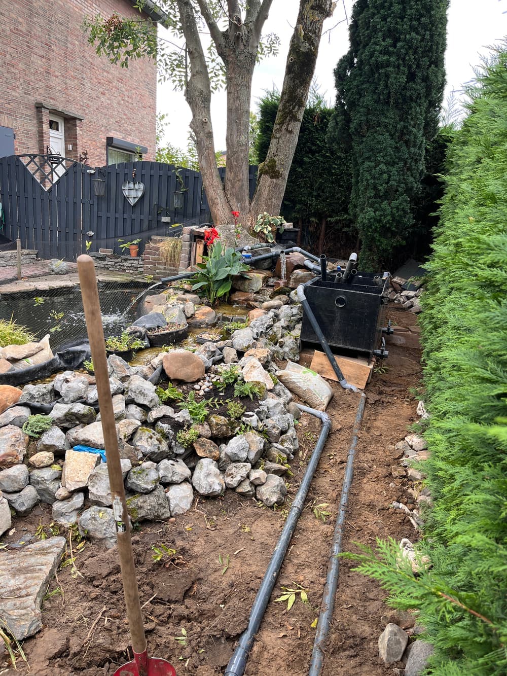 Een gegraven sleuf langs de haag met twee grijze pvc-leidingen, op weg naar het filter naast de vijver.
