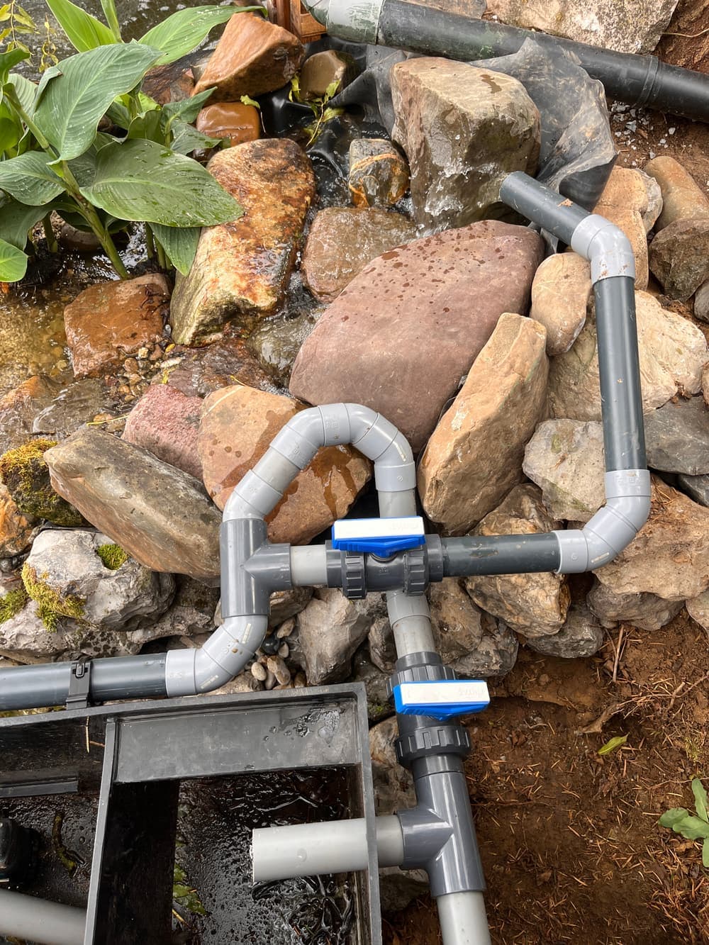Grijze drukvaste pvc-leidingen met twee blauwe kogelkranen tussen de keien bij de waterval.