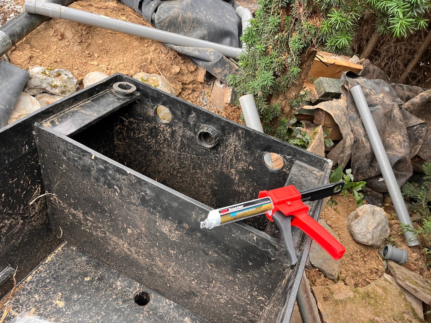 De beschadigde filterbak van dichtbij, met een lijmpistool voor de reparatie en losse pvc-buizen eromheen.