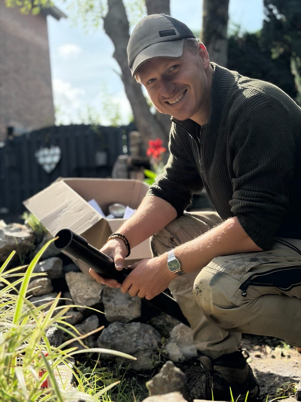 Nick gehurkt tussen de keien aan de vijverrand, lachend, met een zwart onderdeel in de hand en een geopende doos naast zich.